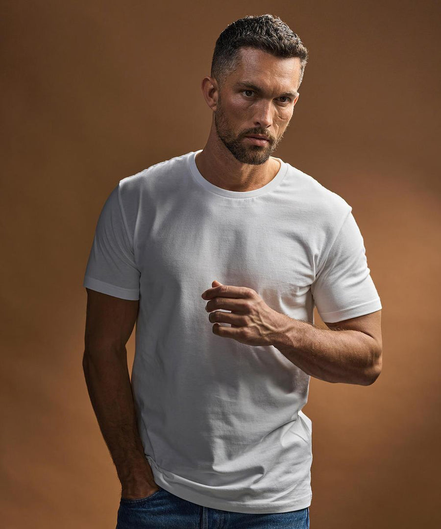 A man with short dark hair and a trimmed beard models the Tee Jays Fashion Sof tee (8005) in white, paired with blue jeans, standing against a brown background with a serious look and one hand raised near his chest.