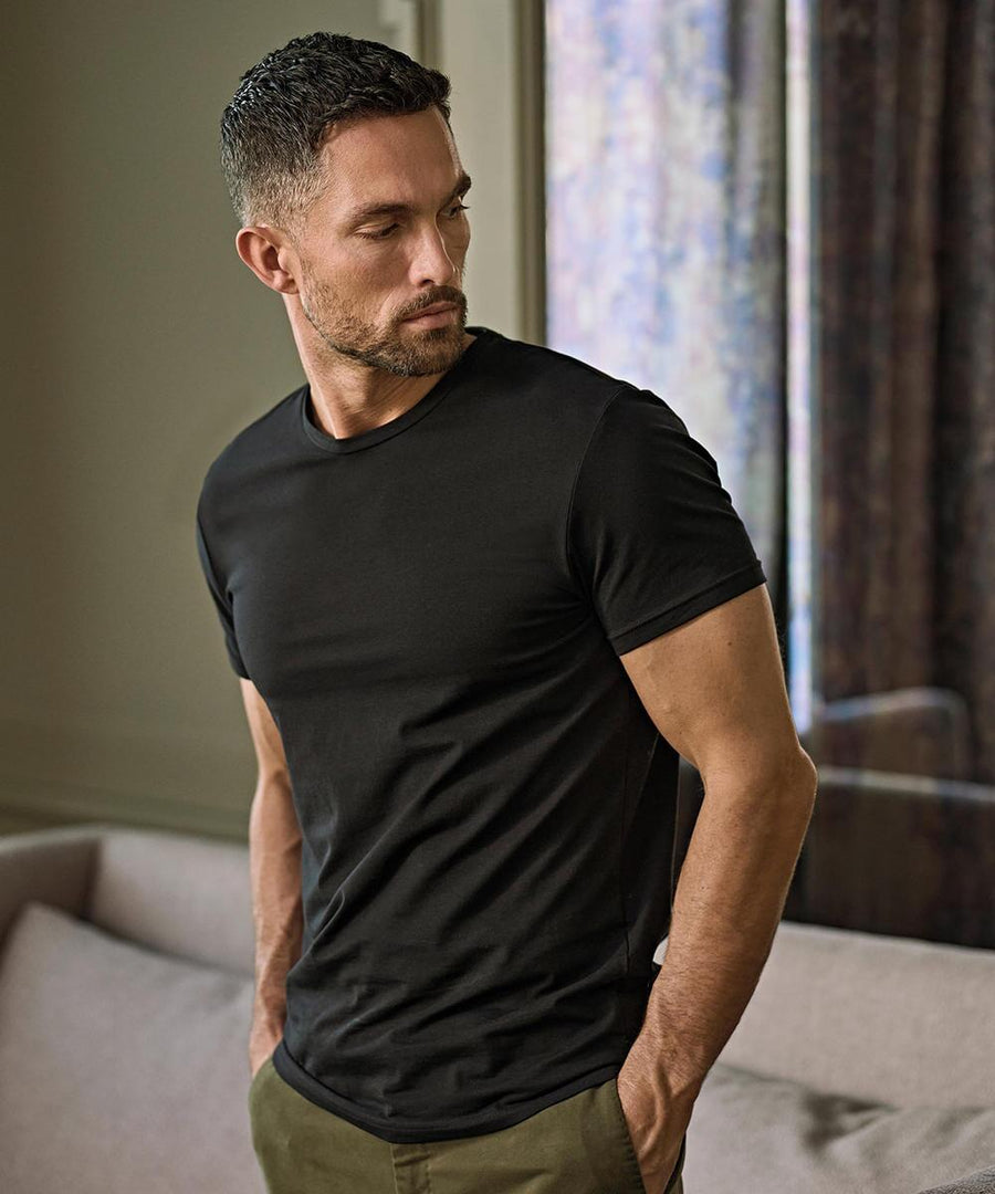 A man with short dark hair and a beard wears the Tee Jays Stretch tee (400) in black and khaki trousers. He stands indoors by a window and light-coloured sofa, looking left with his hands in his pockets.