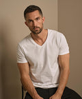 A man with short dark hair and a beard sits on a chair, wearing the Tee Jays Stretch v-neck tee (401) in white and black trousers. He looks directly at the camera against a plain, neutral-toned background.