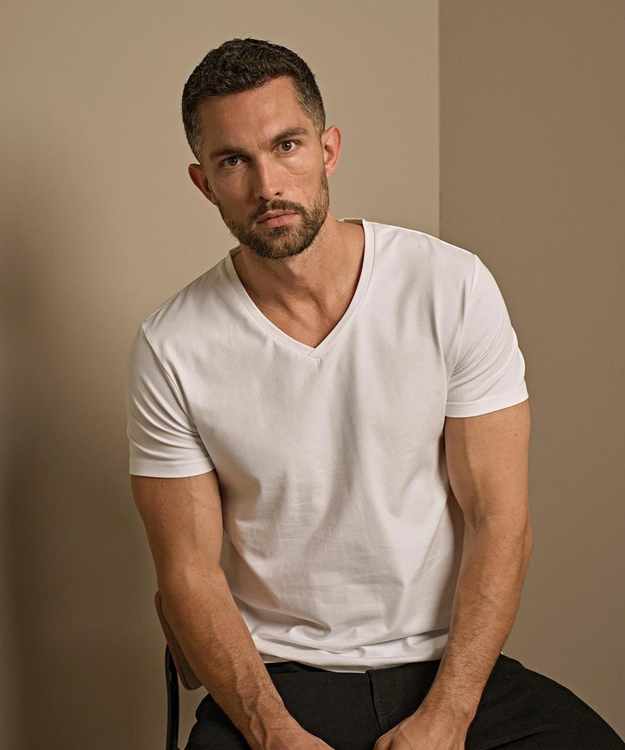 A man with short dark hair and a beard sits on a chair, wearing the Tee Jays Stretch v-neck tee (401) in white and black trousers. He looks directly at the camera against a plain, neutral-toned background.