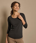 A woman with curly hair wears the Tee Jays Women's stretch 3/4-sleeve tee (460) in black and tan trousers, standing against a beige background, smiling and glancing to the side with one hand touching her collar.