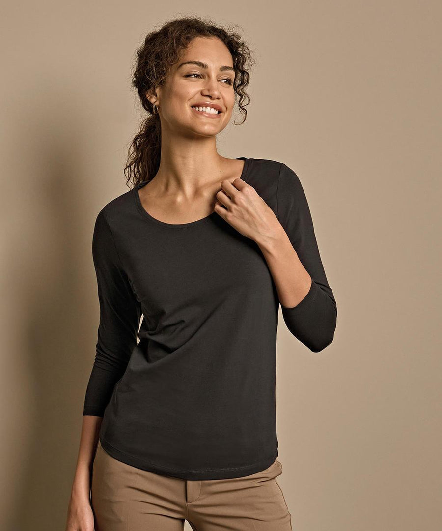 A woman with curly hair wears the Tee Jays Women's stretch 3/4-sleeve tee (460) in black and tan trousers, standing against a beige background, smiling and glancing to the side with one hand touching her collar.