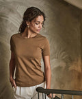 A woman with curly hair stands indoors wearing the Tee Jays Women's interlock tee (580) in brown, paired with white trousers, gold earrings, and a bracelet. She looks down, resting a hand on a metal railing against an artistically textured wall.