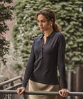 A woman with wavy hair leans on a metal railing outdoors, wearing the Tee Jays Women's luxury stretch long sleeve polo (146) and a light skirt, with evergreen trees in the background.
