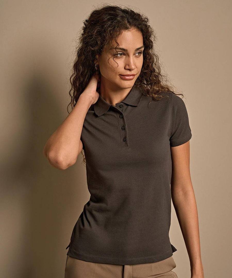 A woman with curly hair models the Tee Jays Women’s heavy polo (1401)—a dark short-sleeve shirt paired with light trousers—standing against a beige background, one hand on her neck and gazing slightly to the side.