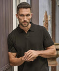 A man with short hair and a trimmed beard stands outdoors, wearing the Tee Jays Luxury stretch v-neck polo (1404) in black, leaning slightly against a wooden wall with his hands clasped in front of him.