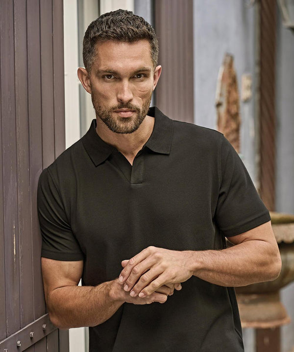 A man with short hair and a trimmed beard stands outdoors, wearing the Tee Jays Luxury stretch v-neck polo (1404) in black, leaning slightly against a wooden wall with his hands clasped in front of him.