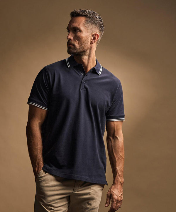 A man with short hair and a trimmed beard stands against a plain brown background, wearing the Tee Jays Luxury Stripe Stretch Polo (1407) in dark navy with white stripe details on the collar and sleeves, paired with light khaki trousers. He looks to his left.