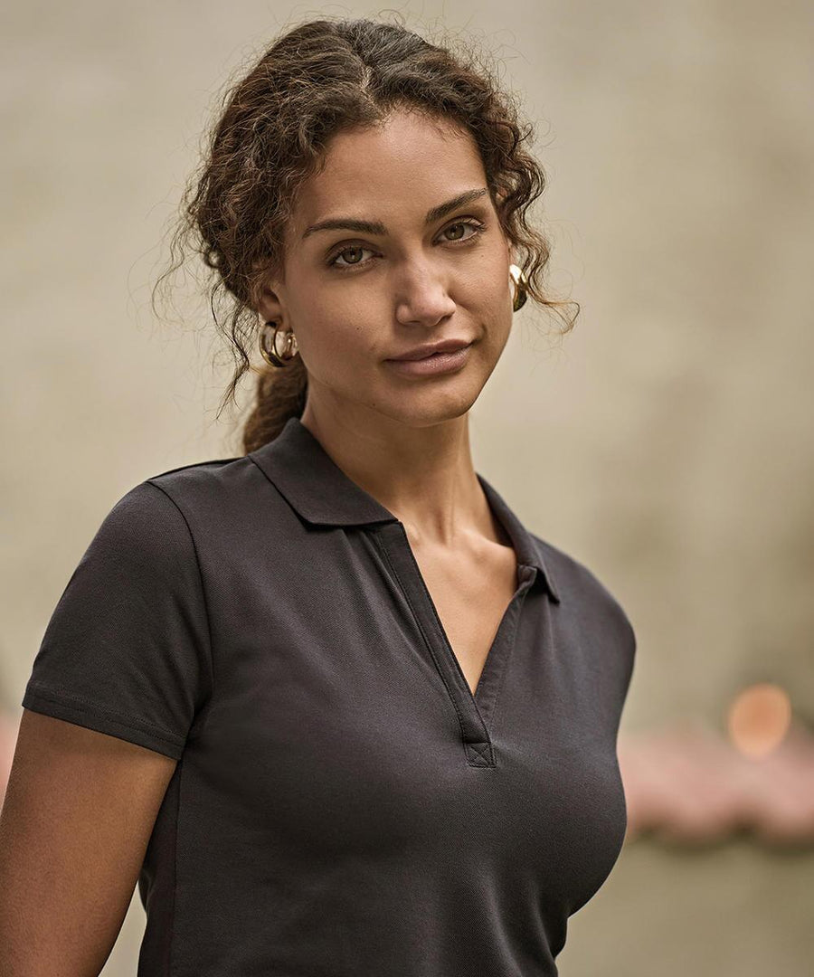 A woman with curly brown hair, wearing the Tee Jays Women’s Luxury Stretch V-Neck Polo (1409) and gold hoop earrings, stands against a blurred neutral background, looking calmly at the camera.