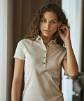 A woman with curly hair stands indoors near a window, touching her ear and looking at the camera whilst wearing a light-coloured, short-sleeved Women's Pima cotton polo (1441) from Tee Jays.