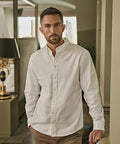A man with short dark hair and a trimmed beard stands indoors, wearing the Tee Jays Perfect Oxford shirt (4000) in white and tan trousers. He looks directly at the camera with a neutral expression; home décor elements appear in the background.