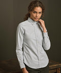 A person with curly hair wears the Tee Jays Women's perfect Oxford shirt (4001) in light grey and dark trousers, standing against a dark background with one hand touching their neck.
