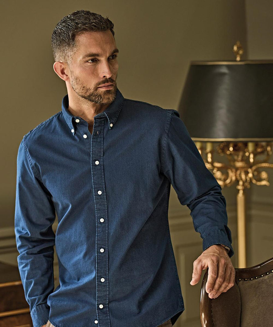A man with short hair and a beard wears the Tee Jays Casual twill shirt (4002) in dark blue, standing beside a chair in a room with gold accents and a lamp. He looks slightly to the side.
