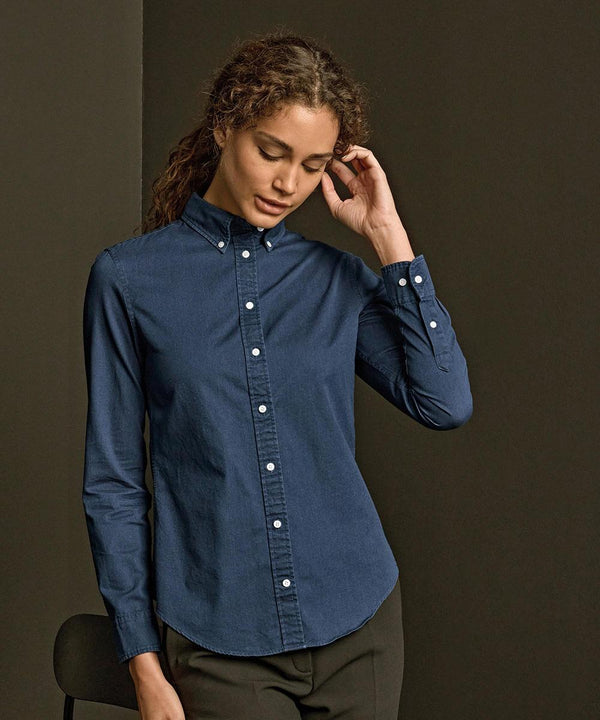 A person with curly hair wearing the Tee Jays Women's casual twill shirt (4003) in dark blue and black trousers stands against a plain dark background, looking down and touching their hair.
