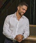 A man with short dark hair and a trimmed beard, wearing a Tee Jays Luxury shirt slim fit (4021) in white and dark trousers, leans against a worktop, smiling as he looks downward and holds his left wrist with his right hand.