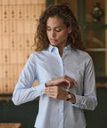 A woman with curly hair, wearing the Tee Jays Women's Stretch Luxury Shirt (4025) in light blue, adjusts her cuff indoors near a wooden table and shelving unit.