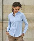 A woman wears the Tee Jays Women’s Active Stretch Shirt (4031), light blue, standing before a stone wall with hands in pockets, looking down with a slight smile.