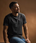 A man with short dark hair and a beard, wearing a Tee Jays Luxury T-shirt (5000) in black and blue jeans, is sitting and looking to his left. The background features a solid brown colour with soft lighting.
