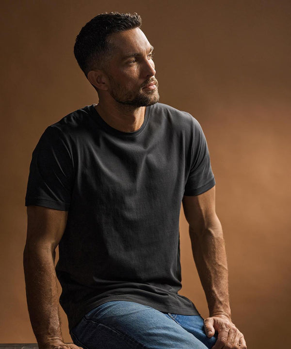 A man with short dark hair and a beard, wearing a Tee Jays Luxury T-shirt (5000) in black and blue jeans, is sitting and looking to his left. The background features a solid brown colour with soft lighting.