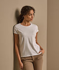 A woman with curly hair stands against a beige wall, wearing the Tee Jays Women’s roll-up tee (5063) in white and light brown trousers. She has one hand in her pocket and gazes to the side with a neutral expression.