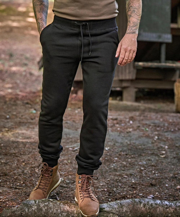 A person stands outdoors on soil, wearing Tee Jays Sweatpants (5425), tan lace-up boots, and a brown shirt. Tattoos are visible on both forearms, with a wooden structure in the background.