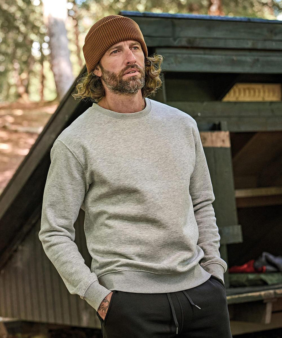 A man with shoulder-length hair and a beard wears a Tee Jays Heavy sweatshirt (5429), along with black trousers and a brown beanie, standing outdoors by a wooden structure with trees in the background.