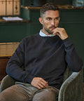 A man with short hair and a beard sits in a chair, wearing the Tee Jays Ribbed Interlock Crew Neck (5504) over a collared shirt and light trousers, resting his elbow on the chair arm and his hand near his mouth, gazing slightly to the side.