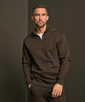 A man with short hair and a beard stands against a dark background, wearing the Tee Jays Ribbed Interlock Half-Zip (5506) pullover and matching trousers. He has a serious expression, with his right hand in his pocket.