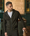 A man with short dark hair and a beard wears the Tee Jays Ribbed Interlock Hooded Full-Zip (5508) in black over a white T-shirt, holding a brown leather bag. He looks to his left in a room with dark green patterned wallpaper.