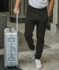 A person in Tee Jays Ribbed interlock trousers (5510), a white t-shirt, and white trainers is walking indoors while pulling a silver rolling suitcase.
