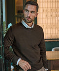 A man with short dark hair and a trimmed beard wears a Tee Jays Crew Neck (6000) jumper over a white collared shirt, standing indoors by a doorway and looking to his left with shelves and a table in the background.