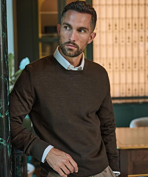 A man with short dark hair and a trimmed beard wears a Tee Jays Crew Neck (6000) jumper over a white collared shirt, standing indoors by a doorway and looking to his left with shelves and a table in the background.
