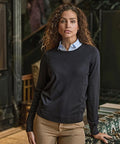 A woman with curly hair stands indoors, wearing the Tee Jays Women's crew neck (6006) in navy blue over a light blue collared shirt and tan trousers. She looks at the camera, set against patterned furniture and dark walls.