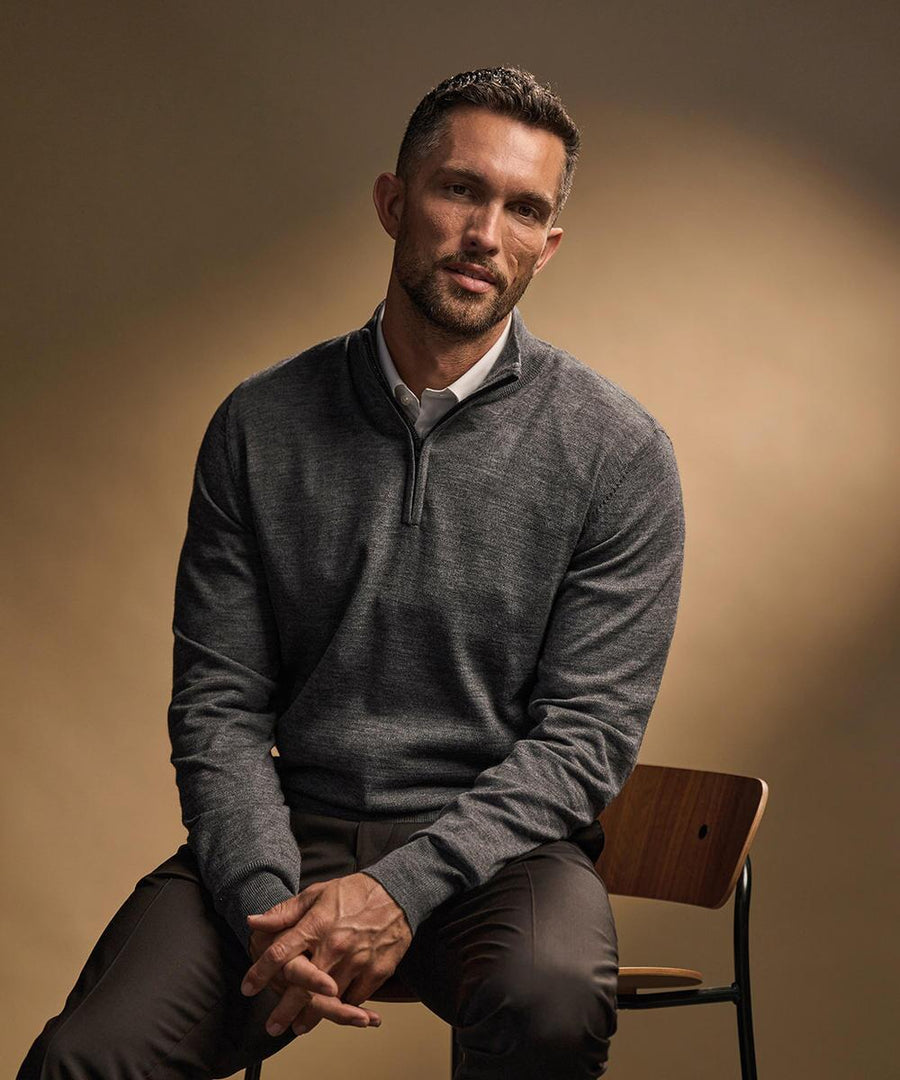 A man with short dark hair and a trimmed beard sits on a wooden chair, wearing the Tee Jays Half-zip (6010) jumper over a collared shirt and brown trousers, looking slightly off camera with a neutral expression.