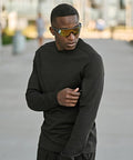 A man wearing mirrored sunglasses and a black Tee Jays Athletic crew neck sweatshirt (5700) stands outdoors, looking to the side as he reaches into a small zip pocket on his sleeve. The background is slightly blurred.
