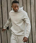 A man wearing a Tee Jays Athletic hooded sweatshirt (5702) and matching tracksuit bottoms stands outdoors by a wooden fence, looking down to his right.