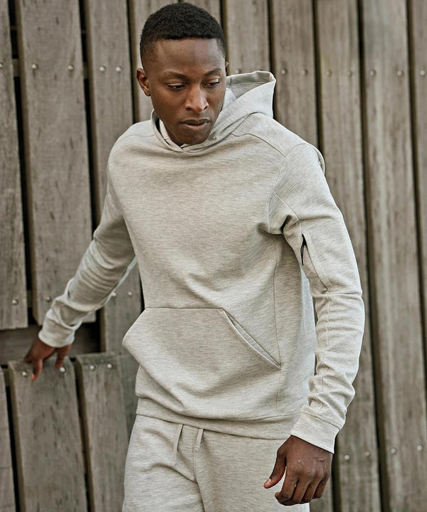 A man wearing a Tee Jays Athletic hooded sweatshirt (5702) and matching tracksuit bottoms stands outdoors by a wooden fence, looking down to his right.
