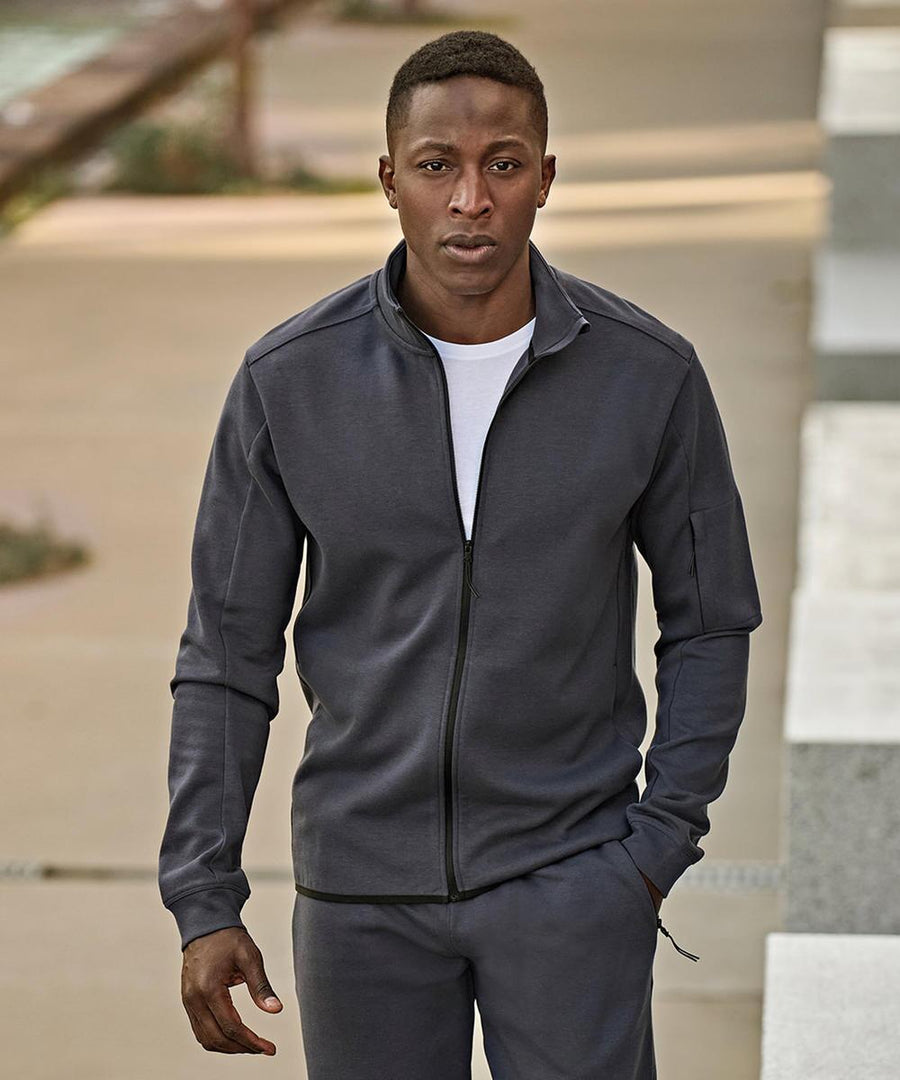 A man stands outdoors in a Tee Jays Athletic full-zip sweatshirt (5704) over a white T-shirt and dark trousers, one hand in his pocket. He has short hair and a neutral expression, with the background slightly out of focus.