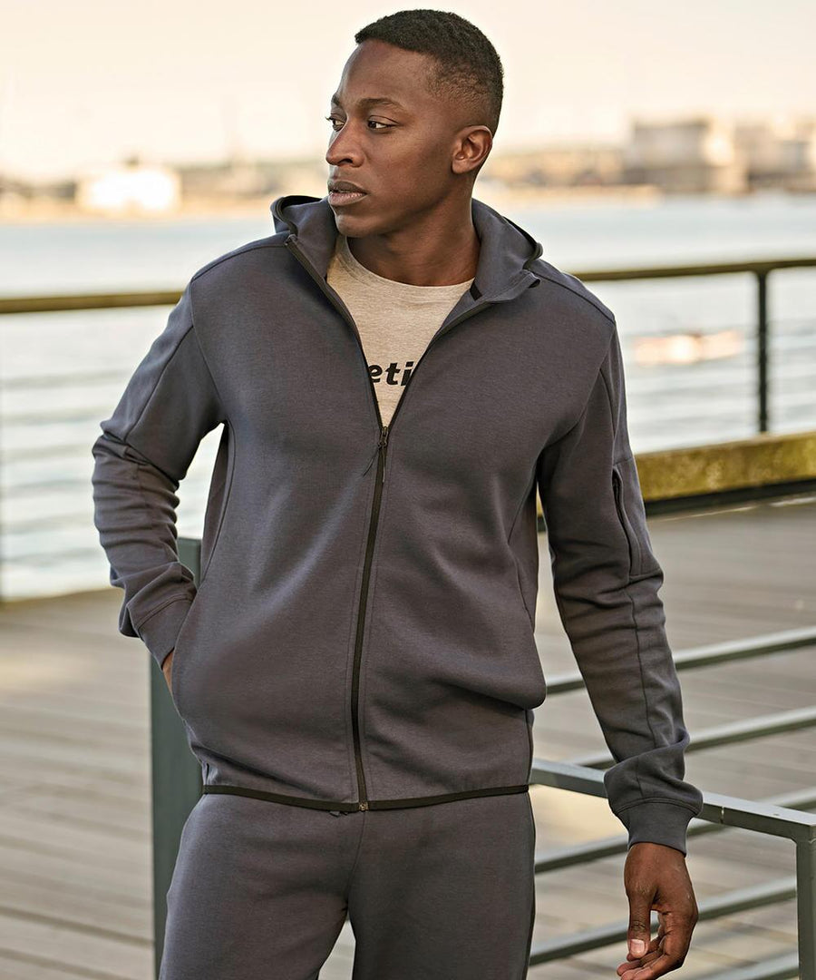 A person wearing the Tee Jays Athletic hooded full-zip sweatshirt (5706) and matching joggers stands on a boardwalk with one hand in their pocket, looking aside. In the background, there’s a body of water and metal railings.