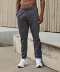 A person stands outdoors in Tee Jays Athletic trousers (5708), dark grey joggers, with hands in pockets and white trainers. The background shows concrete surfaces and vertical wooden panels; the upper body is not fully visible.