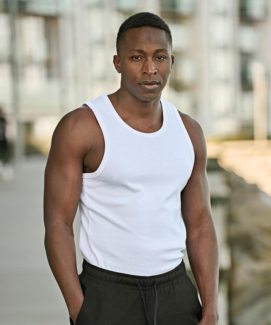 A man with short hair looks directly at the camera outdoors, wearing Tee Jays Athletic shorts (5710) and a white sleeveless shirt. The background is blurred, with buildings and a walkway visible.