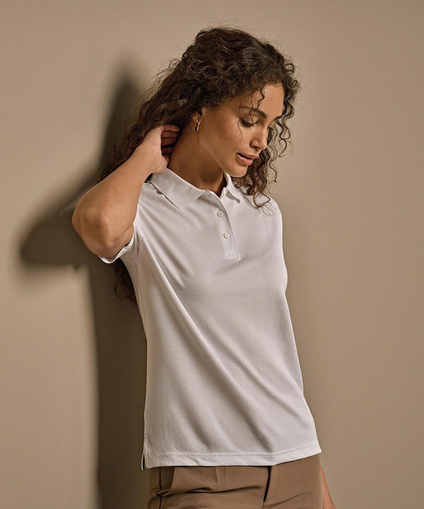 A person with curly hair stands against a beige wall, wearing the Tee Jays Women's Club Polo (7001) in white and tan trousers, with one hand touching the back of their neck and looking downwards.