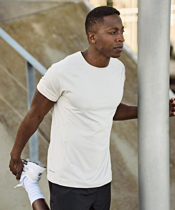 A man wearing a Tee Jays CoolDry tee (7020) and black shorts stretches his left leg outdoors, holding his ankle behind him with his left hand and steadying himself on a pole with his right hand.