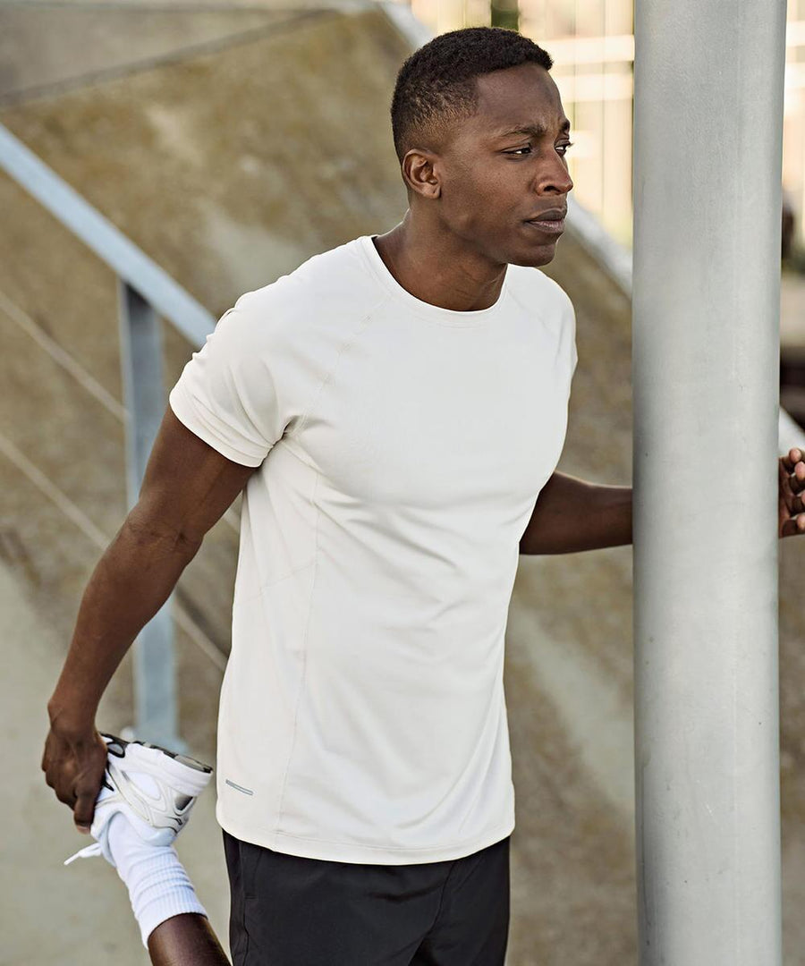 A man wearing a Tee Jays CoolDry tee (7020) and black shorts stretches his left leg outdoors, holding his ankle behind him with his left hand and steadying himself on a pole with his right hand.