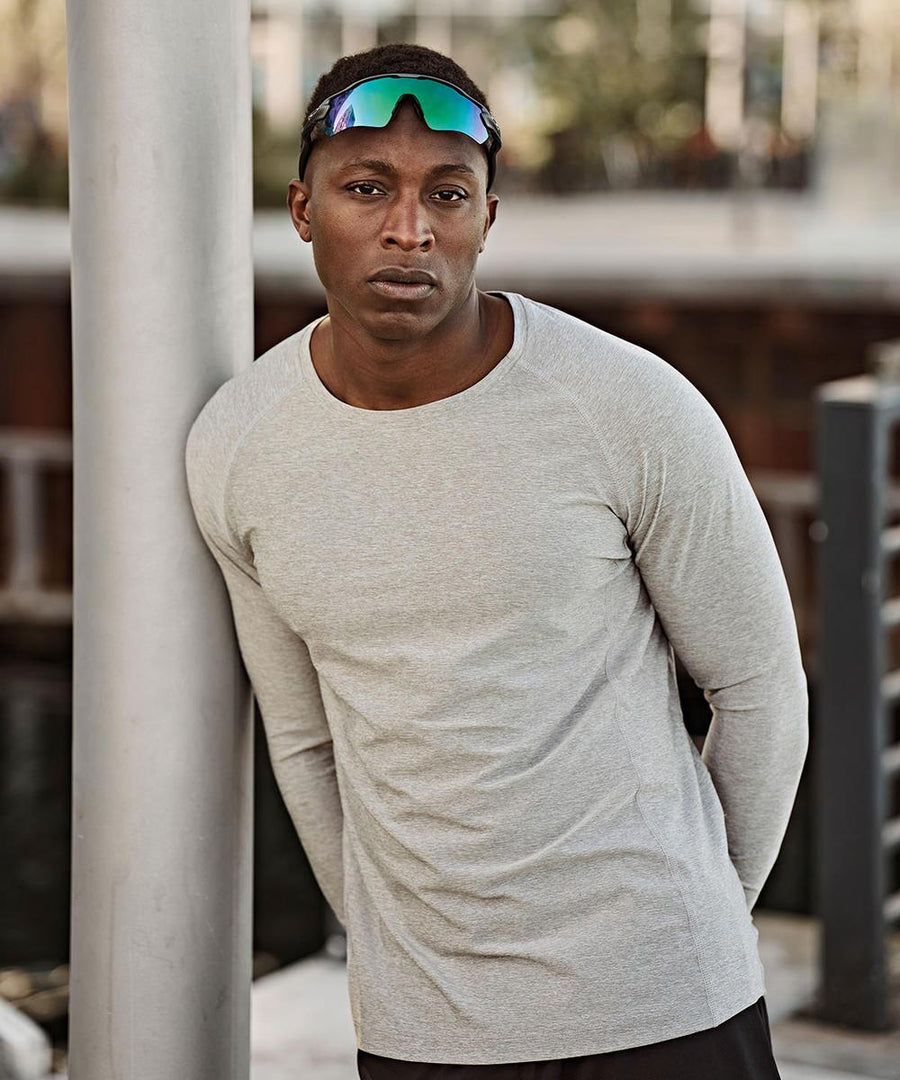 A man wearing a Tee Jays long sleeve CoolDry tee (7022) and reflective sunglasses leans against a pole outdoors, looking at the camera with a neutral expression.