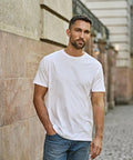 A man with short dark hair and a trimmed beard stands outdoors against a stone wall, wearing the Tee Jays Sof tee (8000) white T-shirt and blue jeans, with one hand in his pocket, looking directly at the camera.