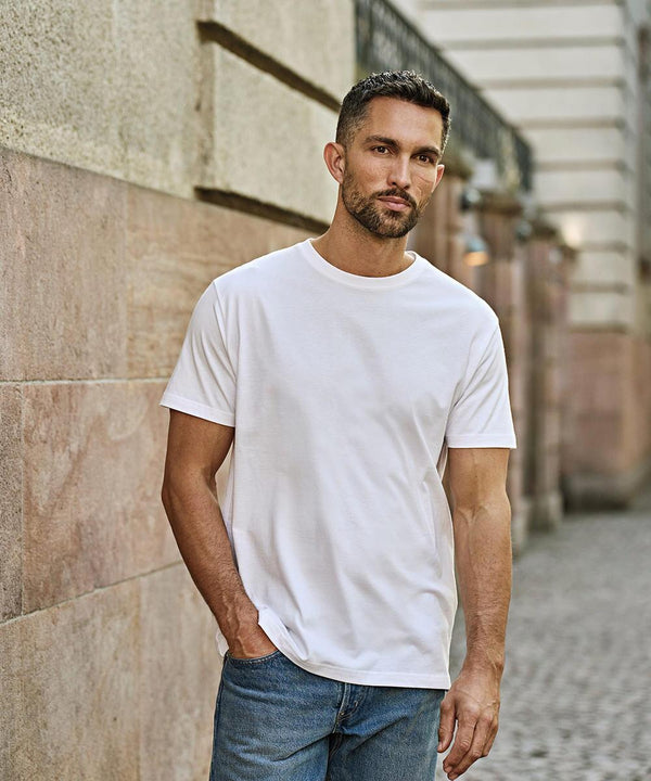 A man with short dark hair and a trimmed beard stands outdoors against a stone wall, wearing the Tee Jays Sof tee (8000) white T-shirt and blue jeans, with one hand in his pocket, looking directly at the camera.