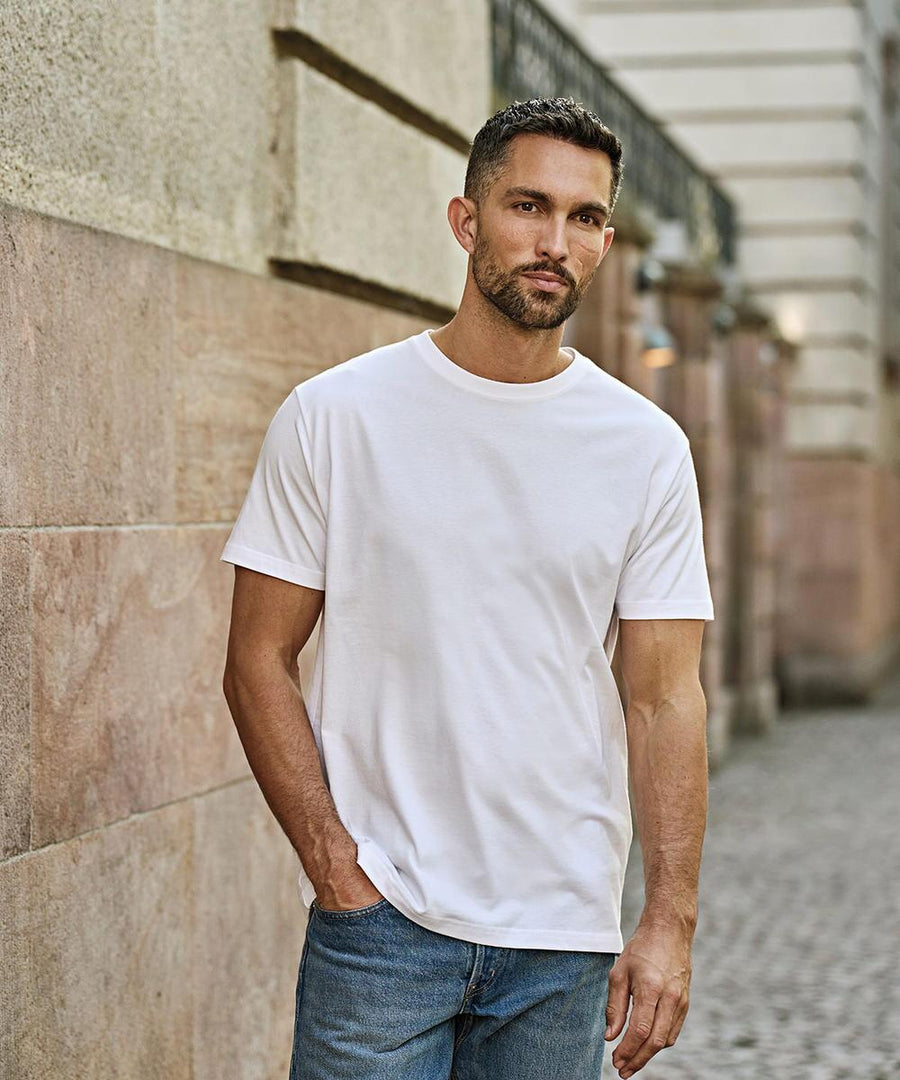 A man with short dark hair and a trimmed beard stands outdoors against a stone wall, wearing the Tee Jays Sof tee (8000) white T-shirt and blue jeans, with one hand in his pocket, looking directly at the camera.
