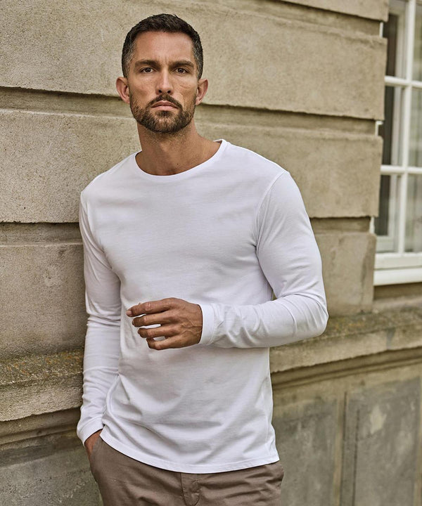 A man with short hair and a beard wears a white Tee Jays long sleeve fashion Sof tee (8007) and light trousers, standing against a stone building, looking at the camera while holding something in his right hand.