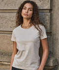 A woman with long curly hair stands against a stone wall, wearing the Tee Jays Womens Sof tee (8050) in a light colour and dark trousers, looking directly at the camera with a neutral expression.